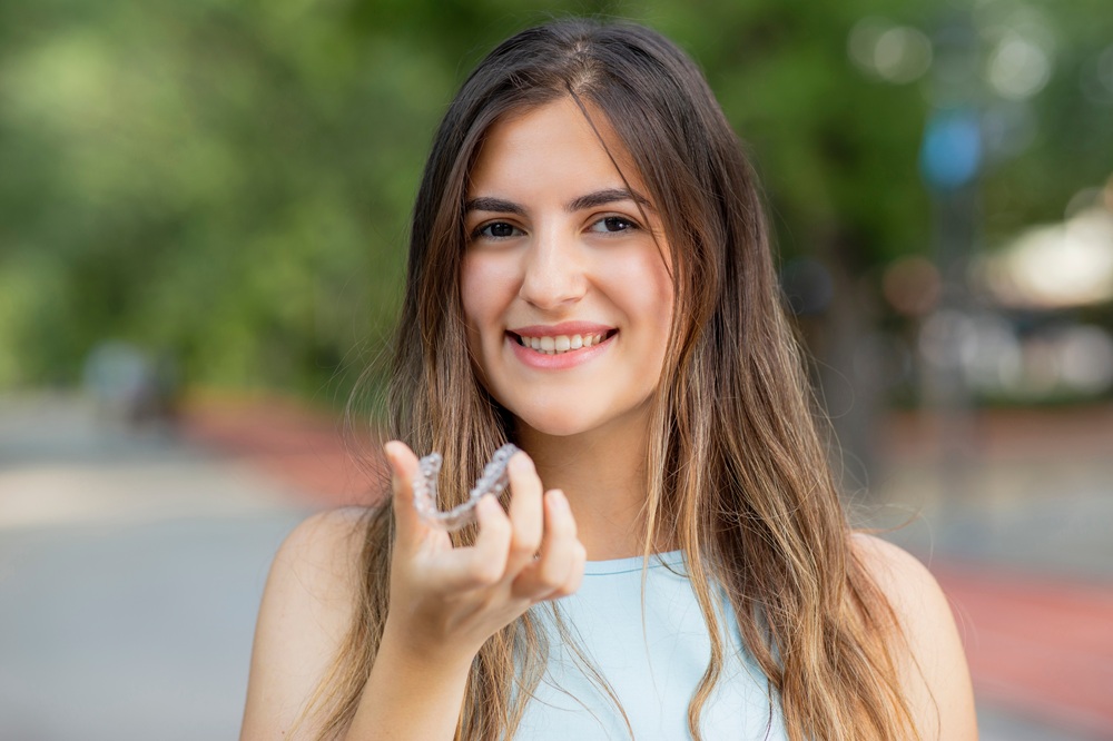 A young woman smiles confidently while holding a clear aligner outdoors, showcasing the discreet and comfortable design of Invisalign treatment - invisalign naples fl