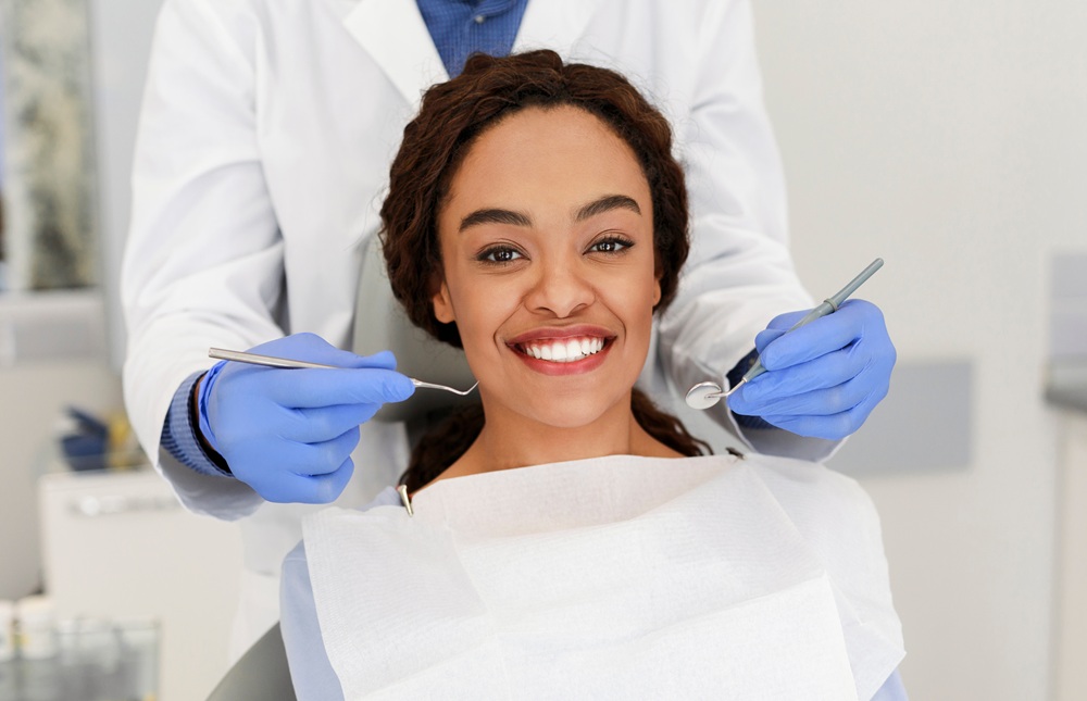 Confident Patient Ready for Dental Treatment - emergency dentist naples A cheerful woman smiles at the camera while sitting in a dental chair as her dentist prepares for an examination with tools in hand - emergency dentist naples