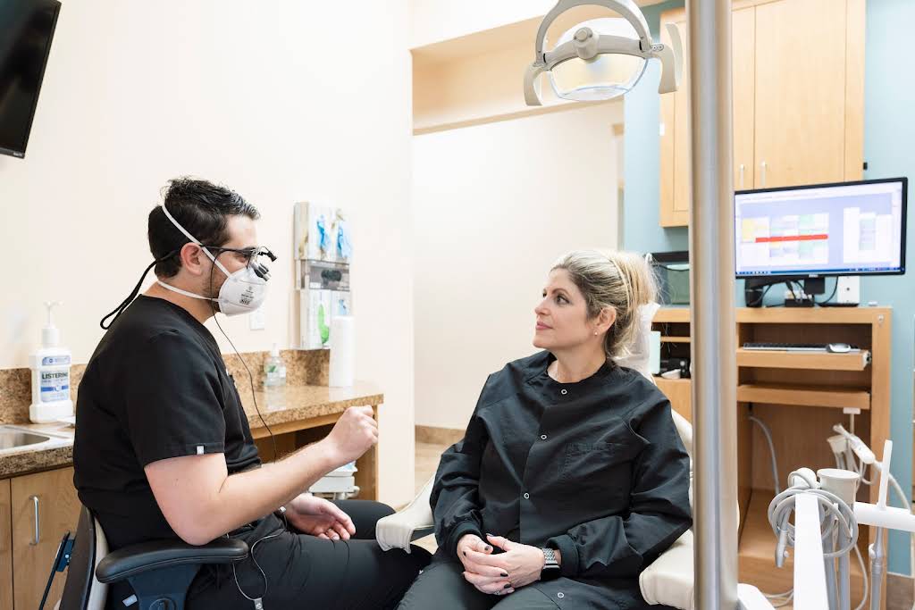 Dentist Consulting with Patient in Modern Dental Office – Dentist in Naples, FL A dentist wearing a mask discusses treatment options with a patient in a bright, modern dental operatory equipped with advanced technology – Dentist in Naples, FL