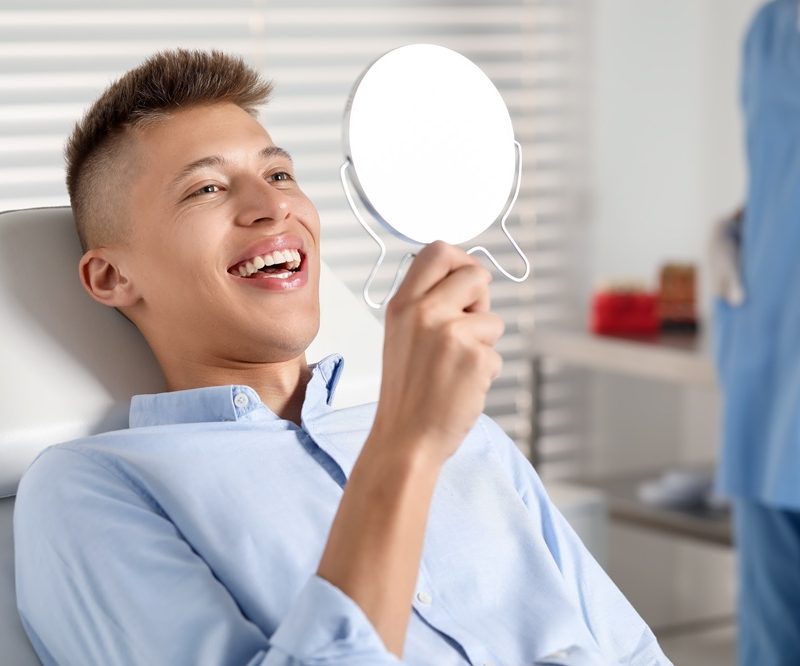 A young man smiles while looking into a handheld mirror during a dental visit, symbolizing satisfaction and confidence after receiving dental crowns - how long does a dental crown last