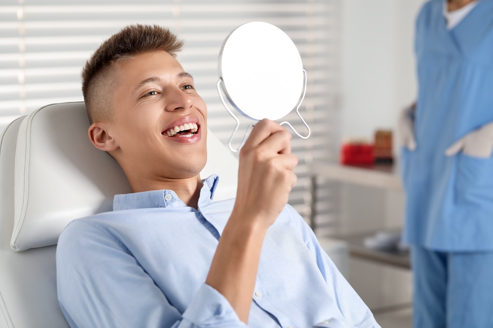 A young man smiles while looking into a handheld mirror during a dental visit, symbolizing satisfaction and confidence after receiving dental crowns - how long does a dental crown last