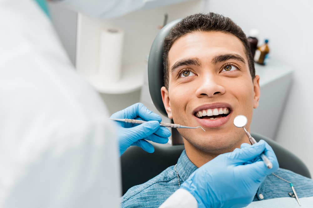Smiling Patient During Dental Exam - emergency dentist naples A young man sits in a dental chair with a bright smile as a dentist performs a checkup using dental instruments and mirror - emergency dentist naples