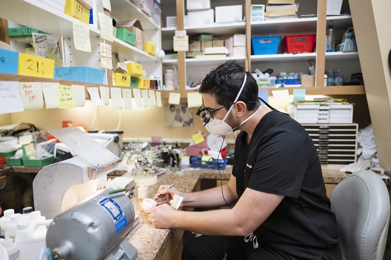 A dental professional wearing magnifying loupes and a mask works on a dental model in a detailed lab setting, showcasing precision and craftsmanship at Suncoast Dental Center.